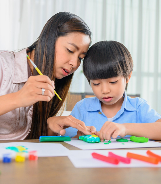 A woman helps a young boy paint and shape colorful clay at a table indoors.