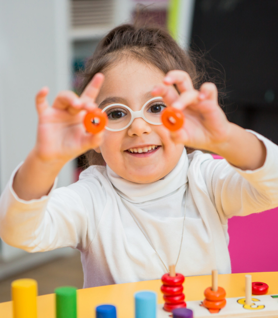 Smiling child wearing white glasses and a white turtleneck holding up orange toy rings while playing at a colorful table.