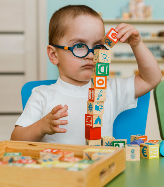 Young boy wearing glasses carefully stacking wooden blocks on a table indoors.