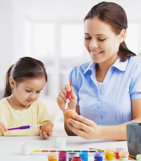 A smiling woman and a young girl painting eggs together at a table with various paint colors and brushes.