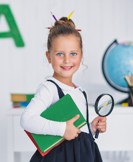 Smiling young girl with pencils in her hair holding a green book and a magnifying glass in a classroom.
