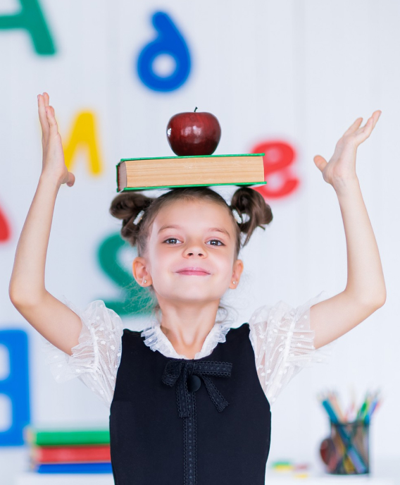 Young girl balancing a green book with a red apple on her head, raising her hands with colorful alphabet letters in the background.