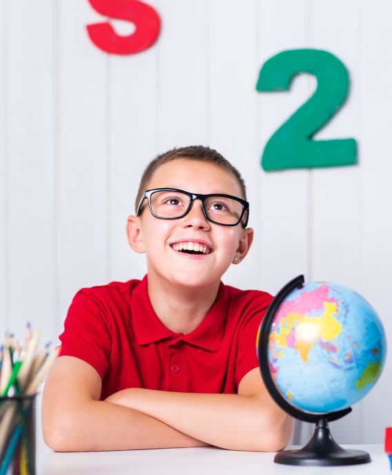Smiling boy with glasses in a red shirt sitting at a desk with a globe and colorful pencils, looking upward.