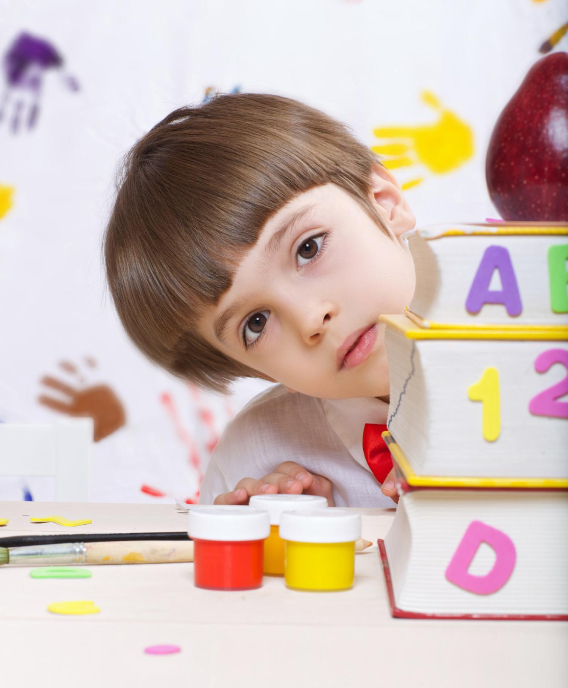 Young boy with brown hair leaning sideways behind a stack of colorful alphabet and number books with paint jars and a red apple nearby.