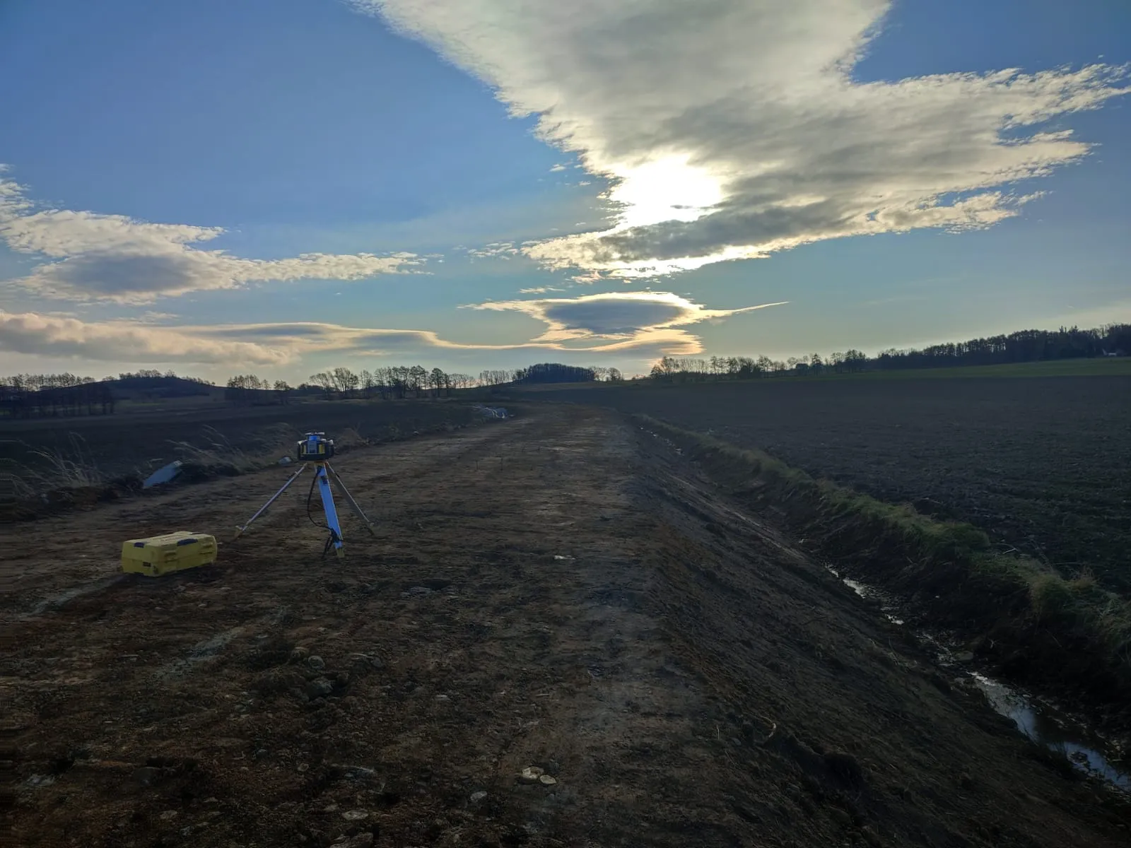 Urządzenie geodezyjne na statywie na placu budowy z polami i niebem z chmurami o zachodzie słońca w tle.