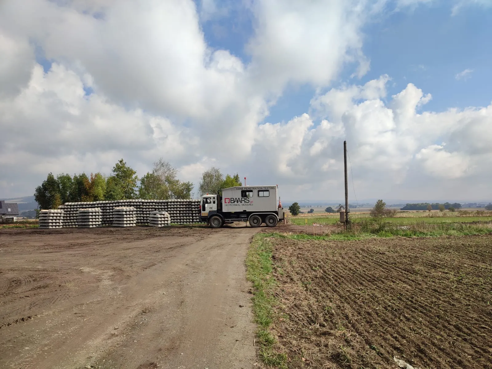 Ciężarówka do badań geotechnicznych obok ułożonych betonowych elementów na terenie budowy na wsi pod chmurzystym niebem.