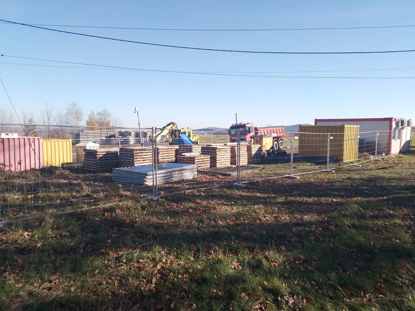 Plac budowy z materiałami budowlanymi, maszyną koparką, czerwonym wywrotką i kontenerami magazynowymi otoczony metalowym ogrodzeniem.