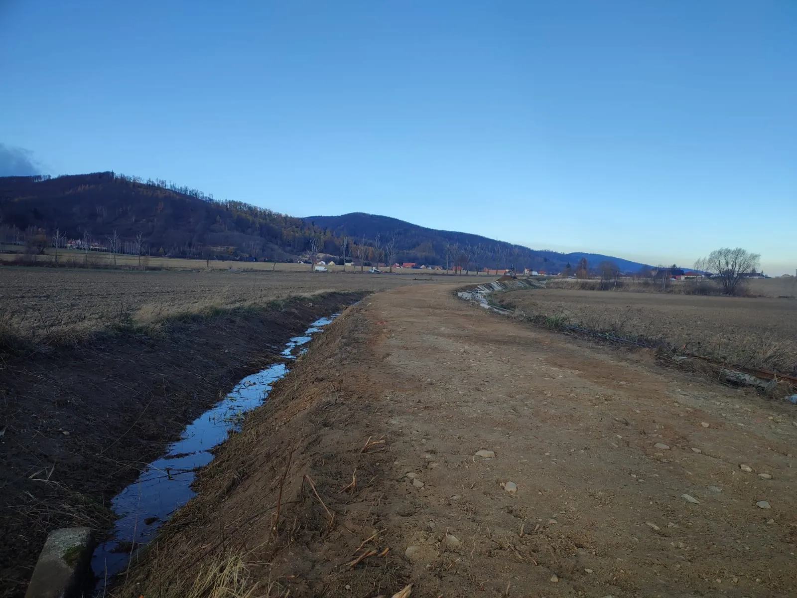 Błotnista droga obok kanału z wodą, z polami i wzgórzami w tle pod błękitnym niebem.