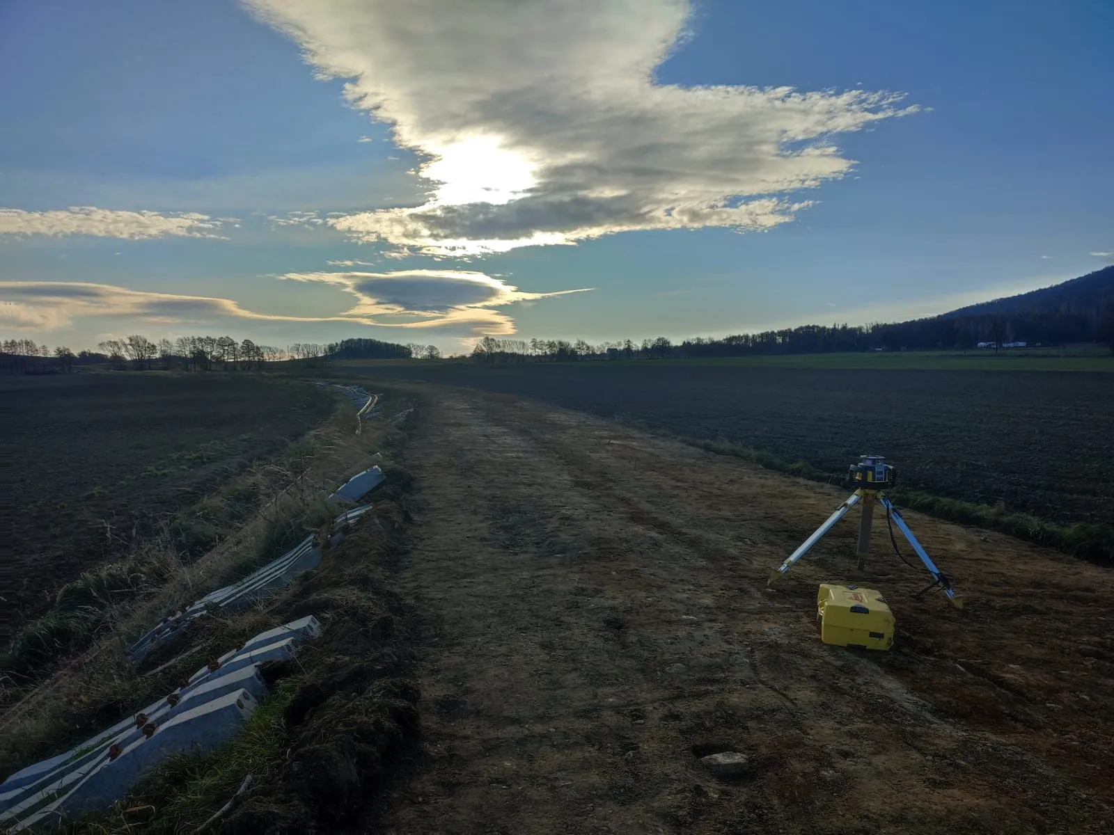 Pusty teren budowy z urządzeniem pomiarowym na statywie i skrzynką na ziemi przy drodze.