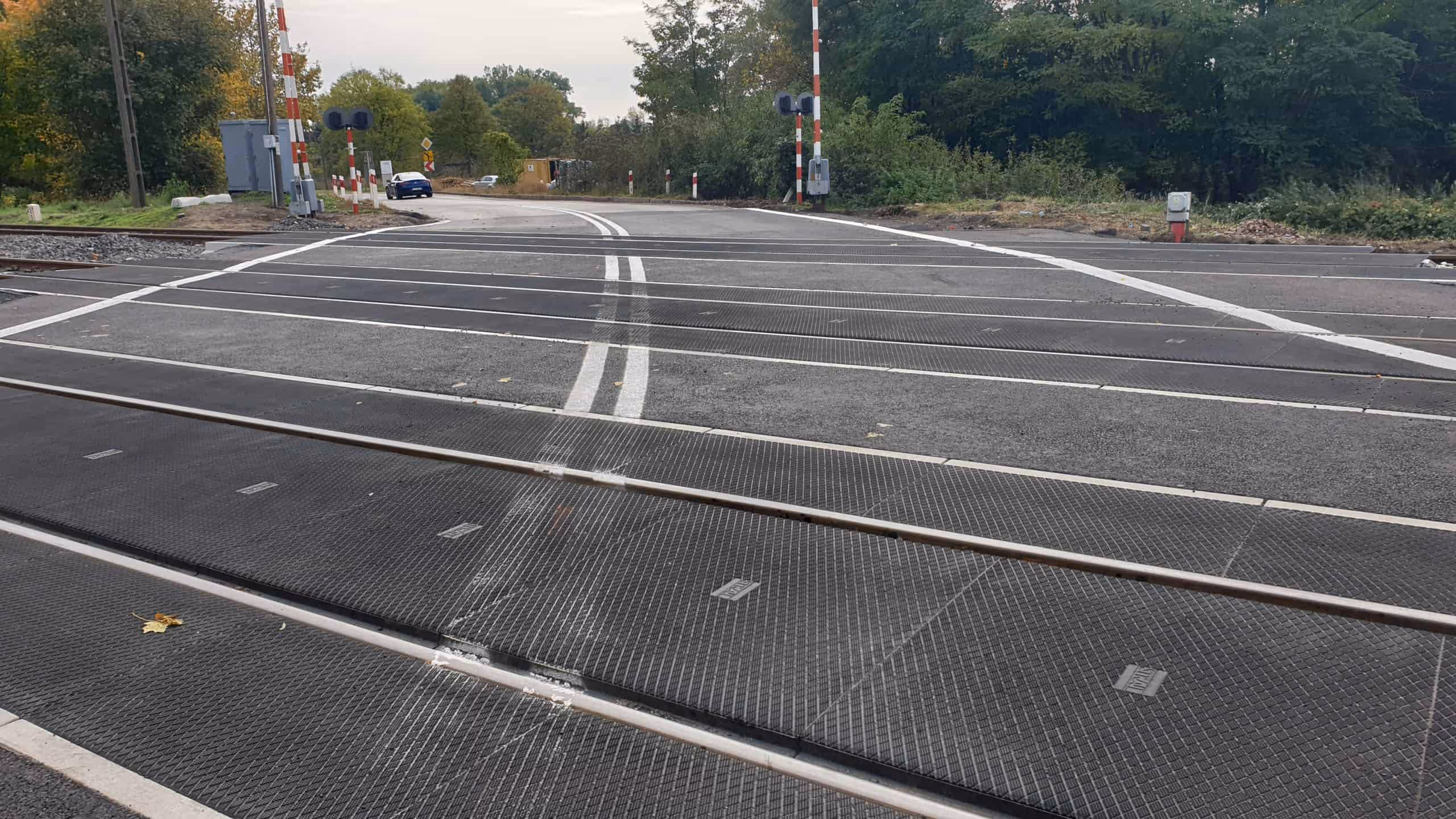 Przejazd kolejowy z czterema szynami i zaporami po obu stronach, otoczony zielenią i drogą asfaltową z liniami malowanymi na jezdni.