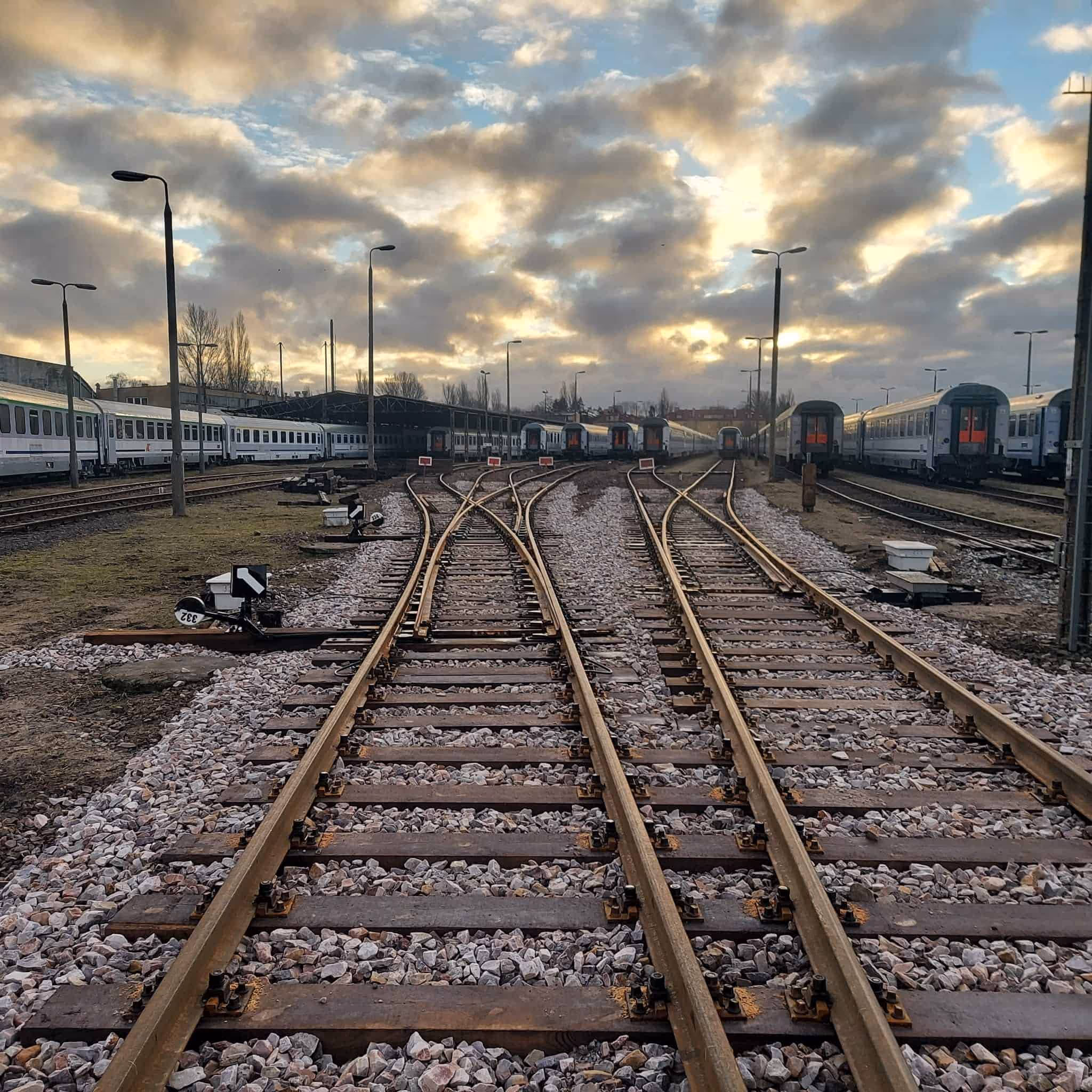 Tory kolejowe na stacji z zaparkowanymi pociągami pod niebem z chmurami o zachodzie słońca.