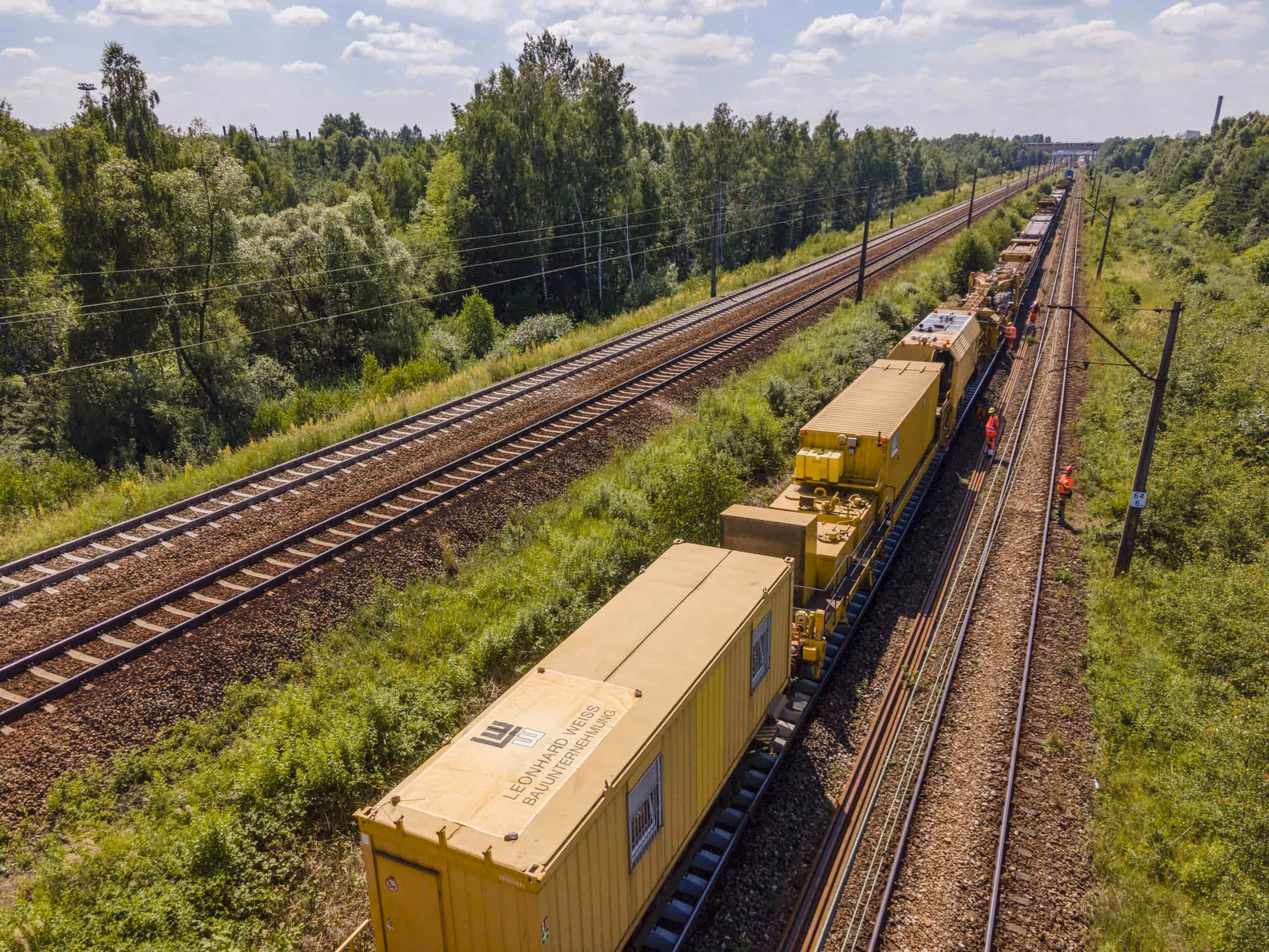 Długi pociąg roboczy na torach kolejowych otoczonych zielenią i lasem, z kilkoma pracownikami w kamizelkach na torach.