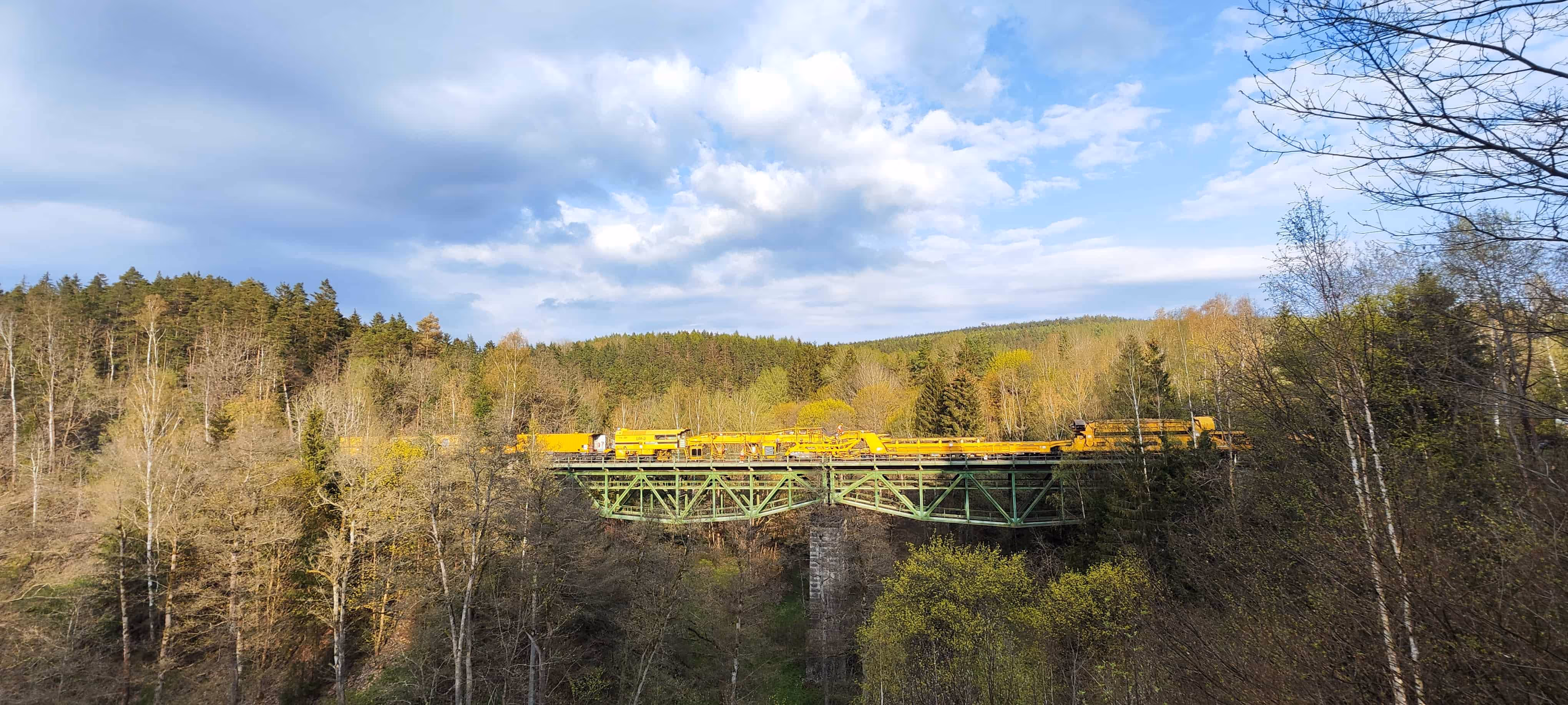 Żółty pociąg roboczy na zielonym moście nad lasem pod niebieskim niebem z chmurami.