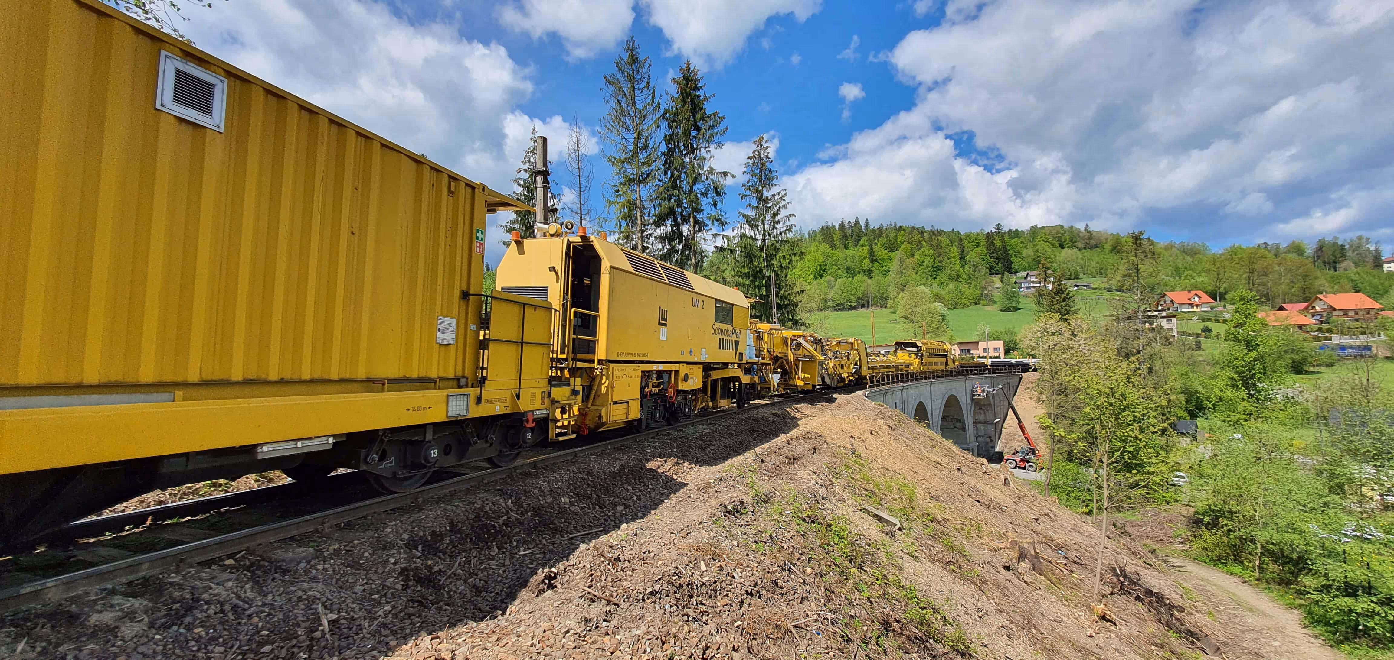 Żółty pociąg roboczy na torach kolejowych przejeżdżający przez kamienny wiadukt w zielonym, pagórkowatym krajobrazie.