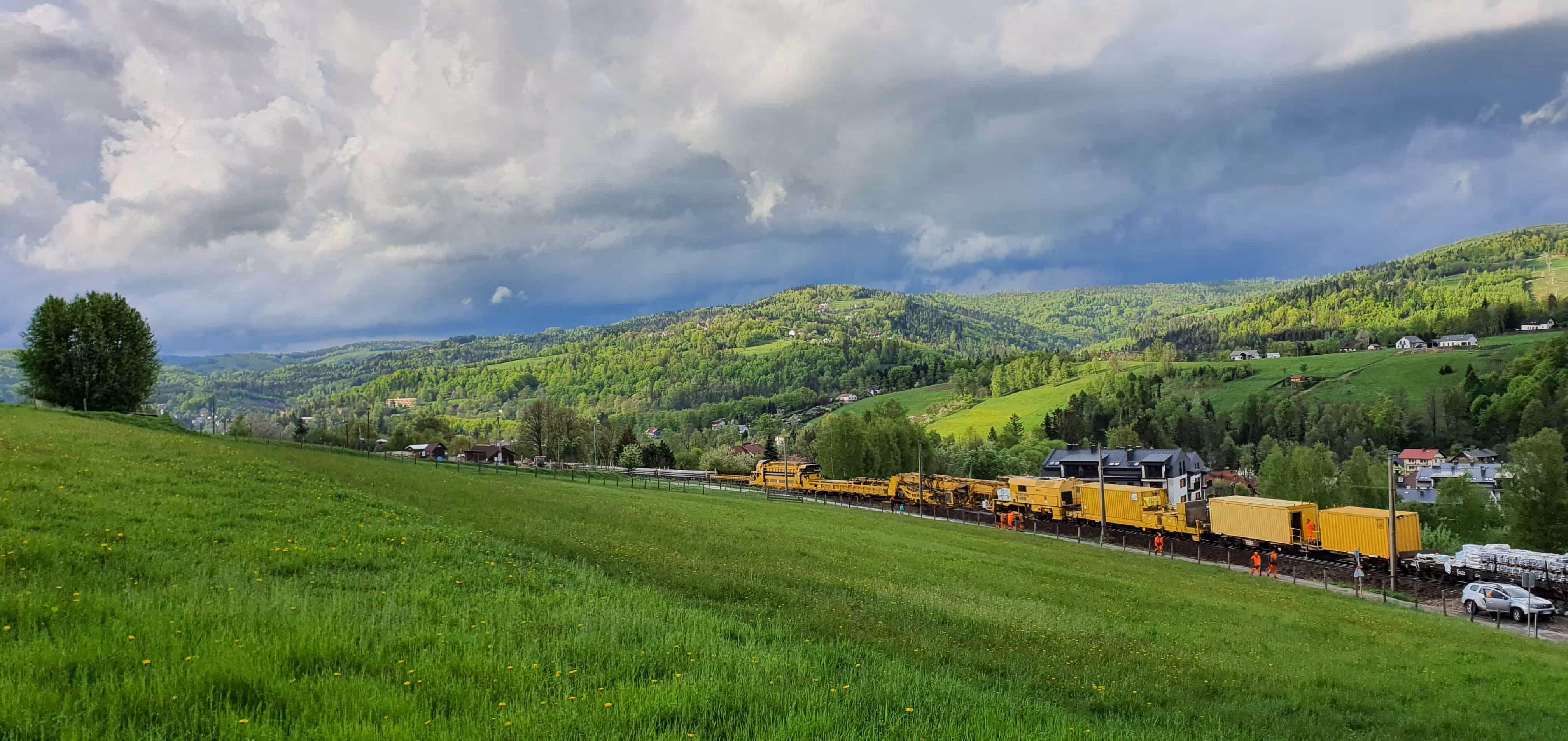 Żółty pociąg roboczy na torach wśród zielonych wzgórz i domów pod zachmurzonym niebem.