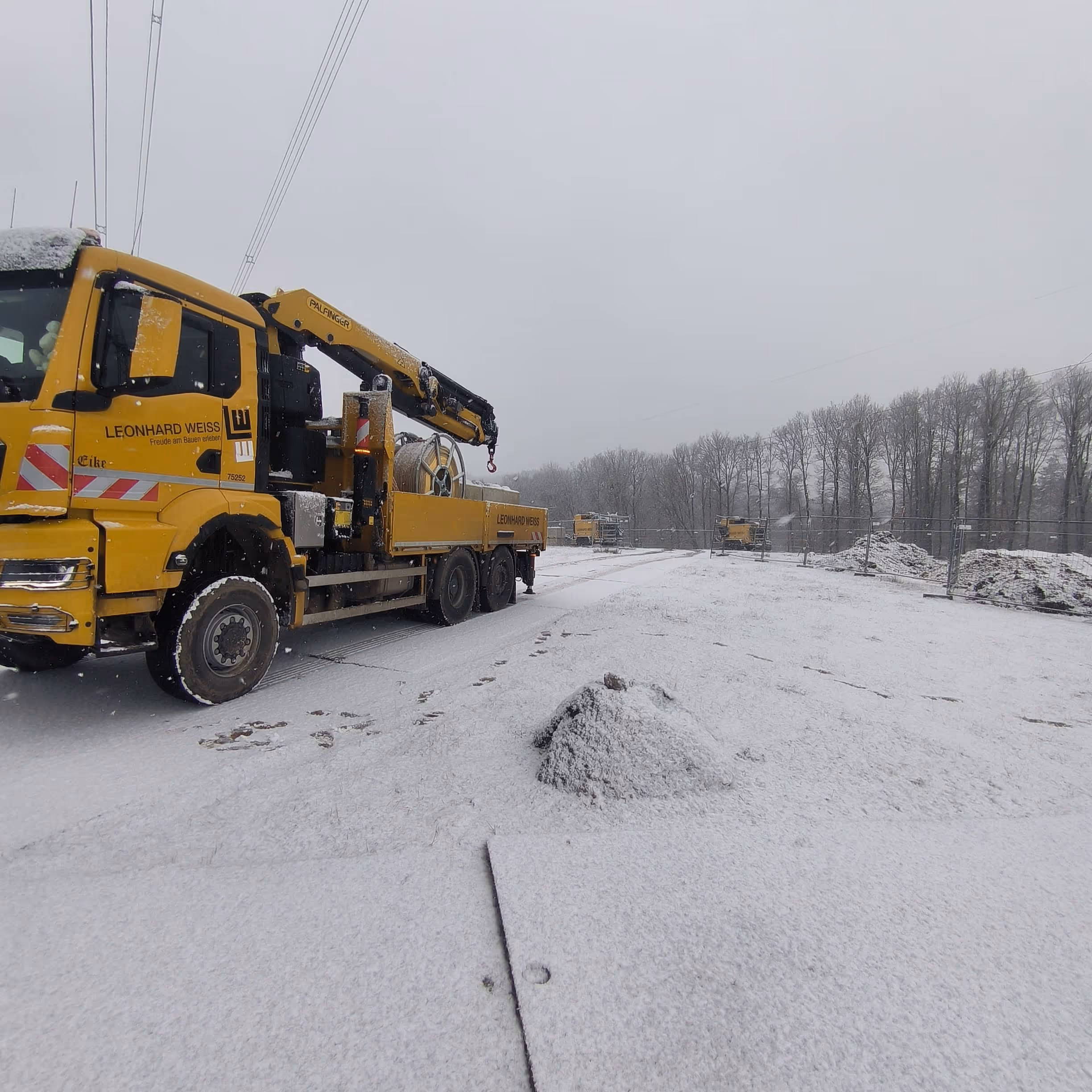 Żółta ciężarówka z dźwigiem na zaśnieżonym placu budowy z drzewami w tle.