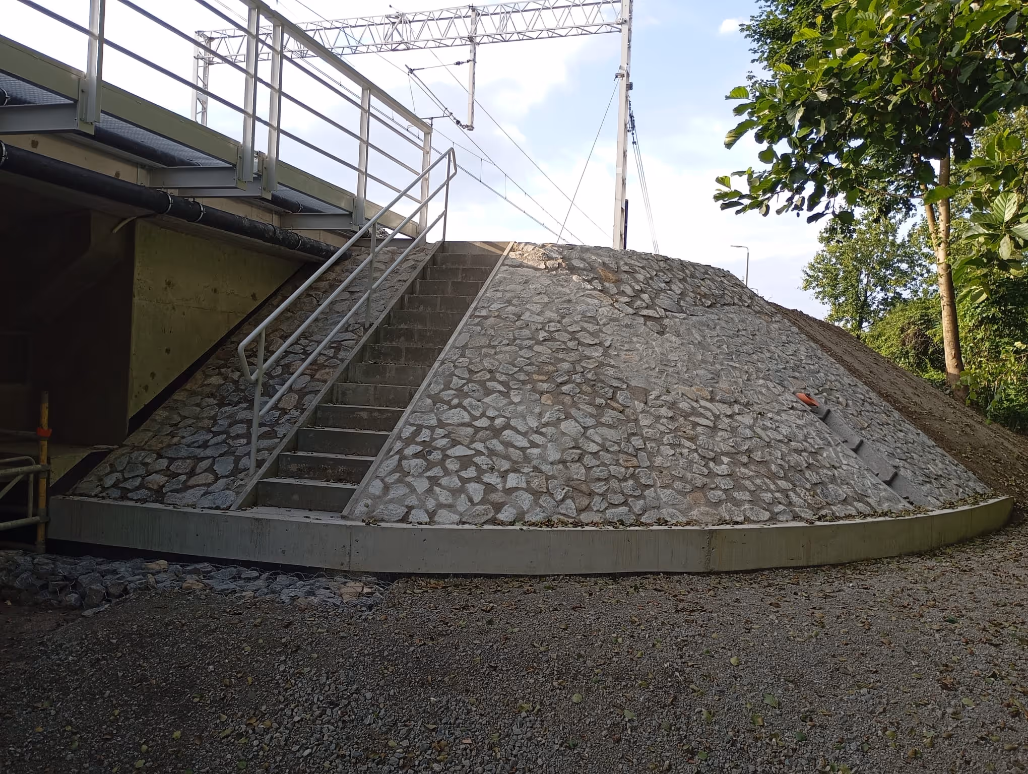 Kamienna skarpa z betonowymi schodami i poręczą prowadzącymi pod most kolejowy z metalową balustradą.