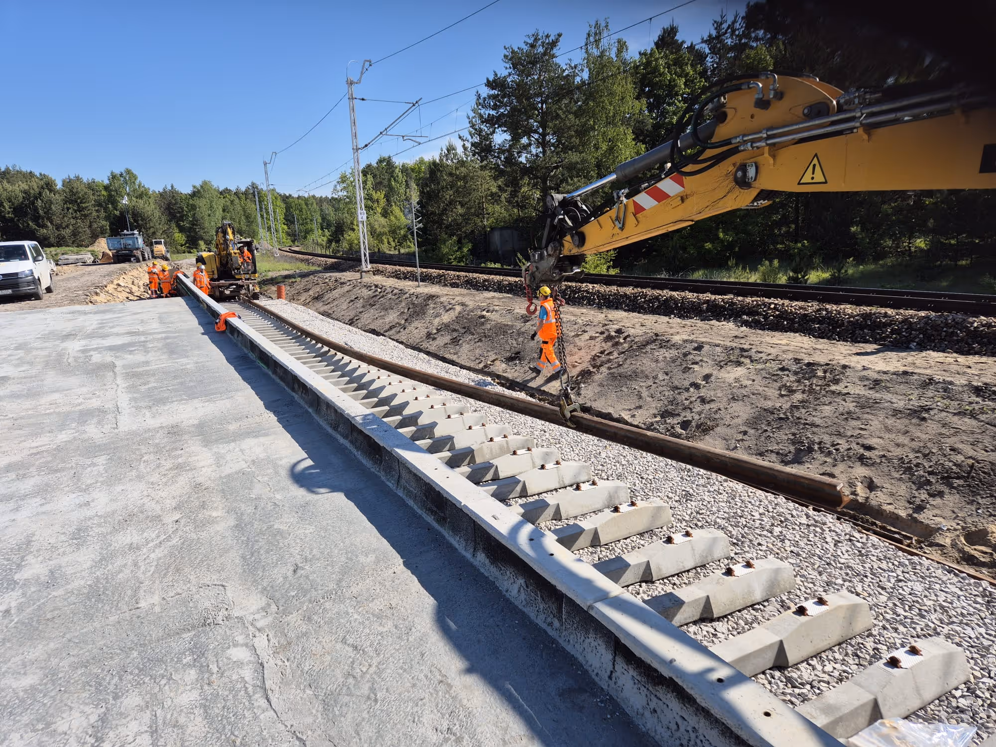 Robotnicy zakładają szyny kolejowe obok istniejącego toru przy użyciu dźwigu i koparki na terenie zalesionym.