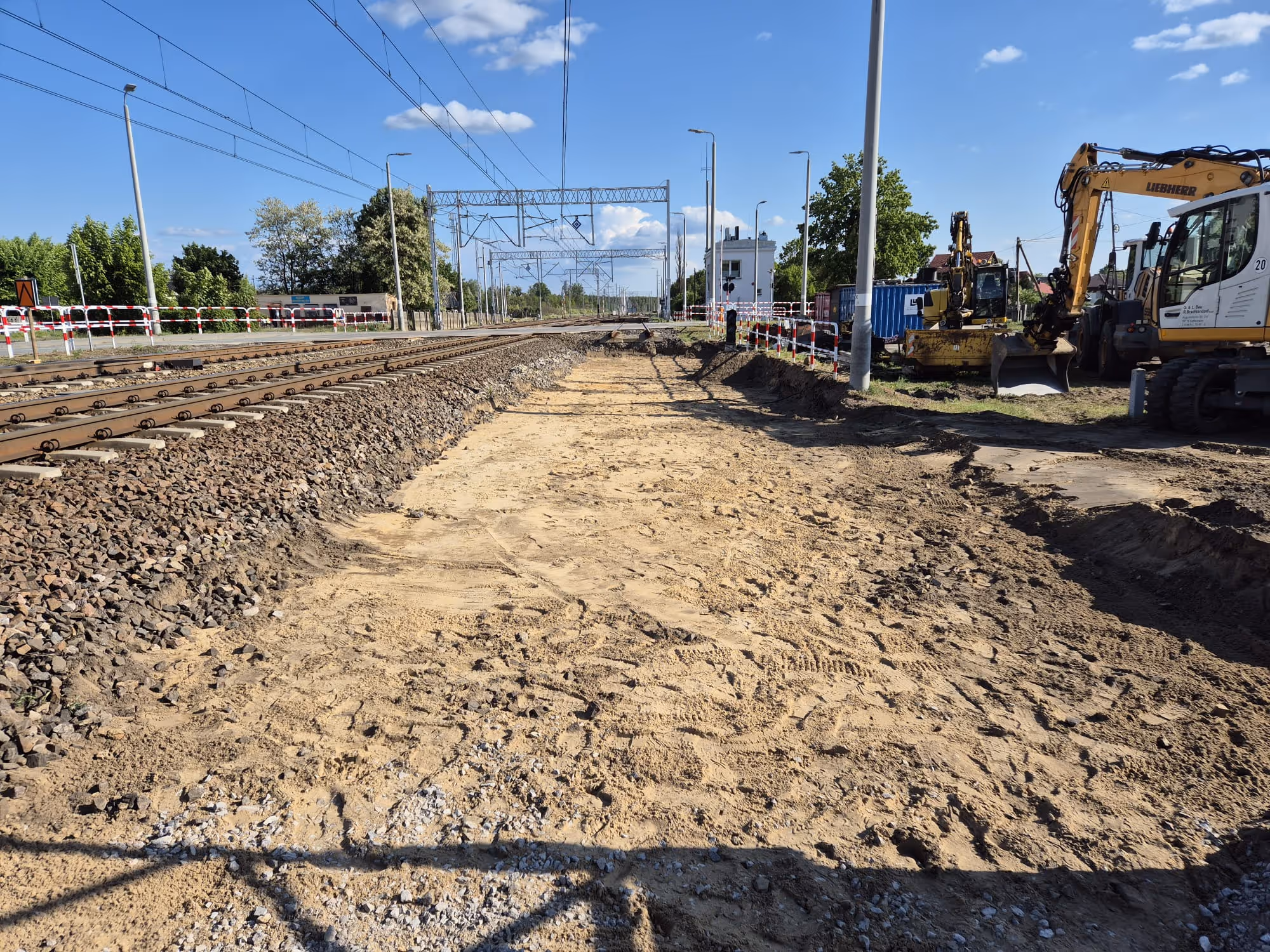 Plac budowy obok torów kolejowych z maszynami budowlanymi i wykopanym terenem.