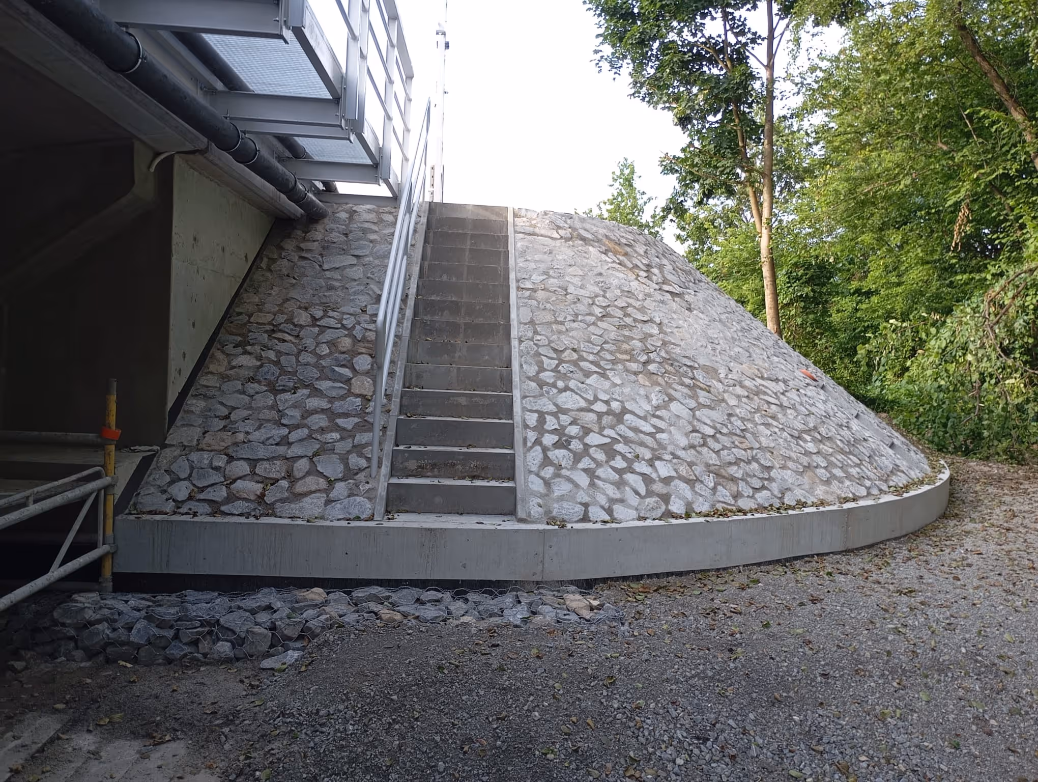 Kamienna skarpa z betonowymi schodami i metalową barierką obok zielonych drzew.