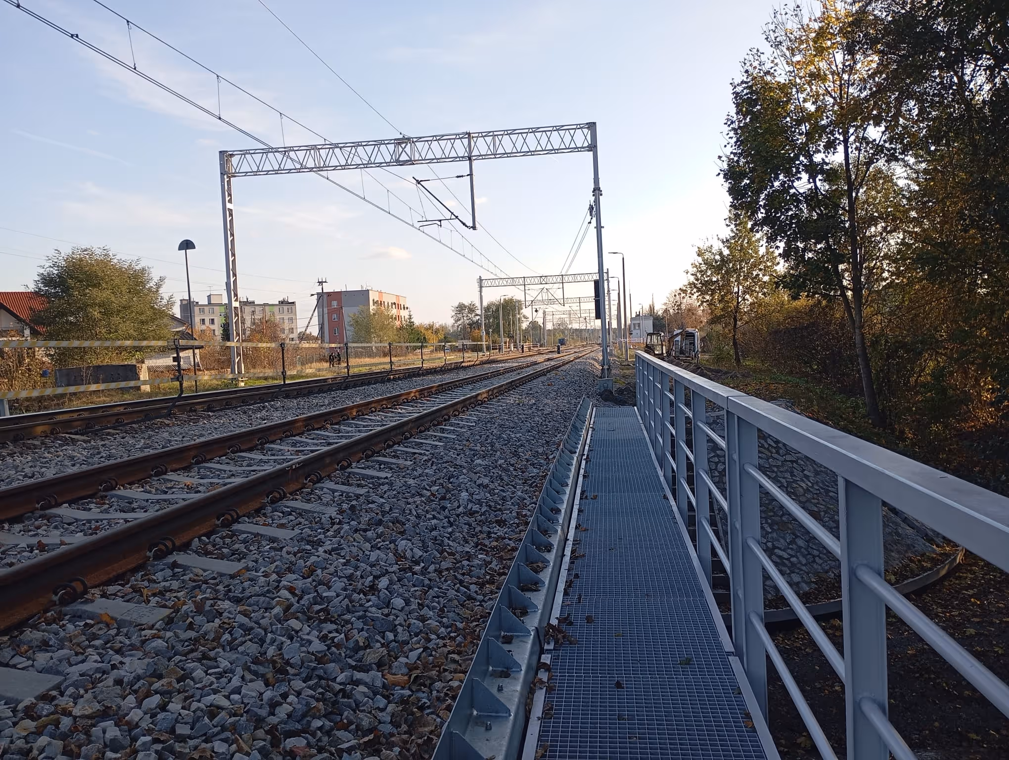 Szyny kolejowe biegnące wzdłuż kamienistej podsypki z chodnikiem i metalową balustradą po prawej stronie, otoczone drzewami i budynkami w tle.