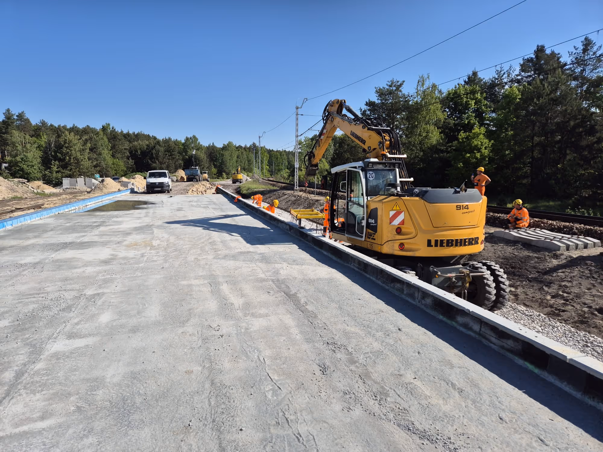 Budowa peronu kolejowego z ciężkim sprzętem i pracownikami w pomarańczowych ubraniach ochronnych.