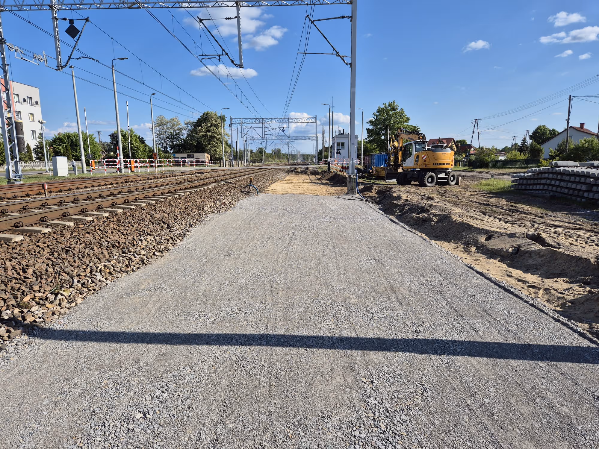 Tory kolejowe i plac budowy z koparką obok torów pod linią trakcyjną na tle niebieskiego nieba.