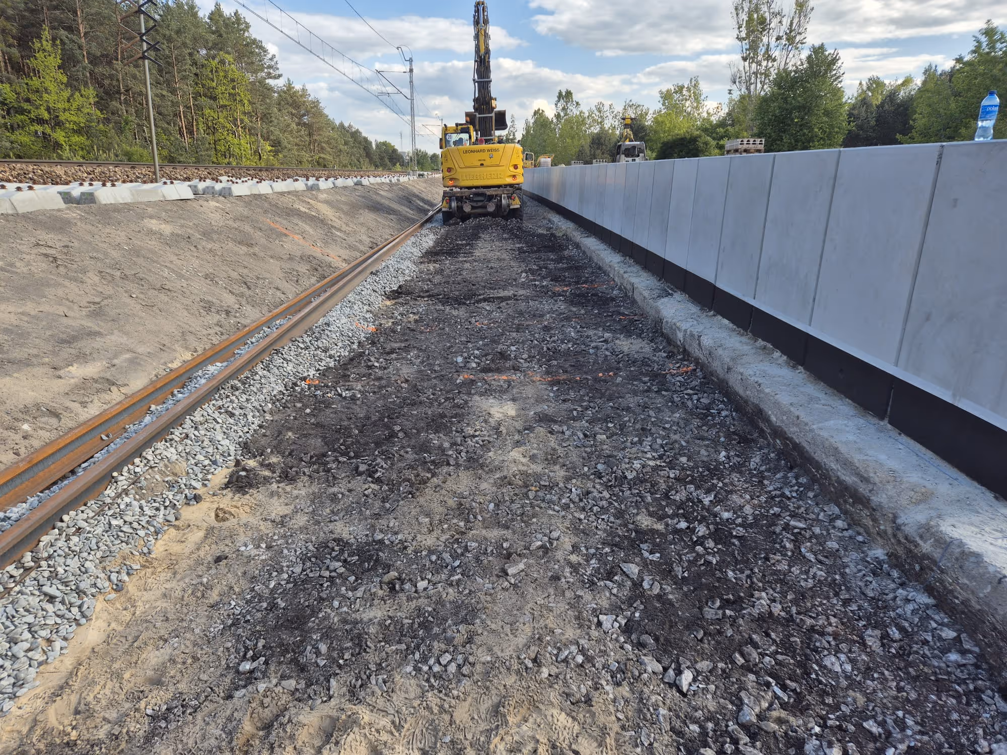Tory kolejowe i teren budowy zabezpieczonej betonową ścianą dźwiękoszczelną, z maszyną do prac torowych.