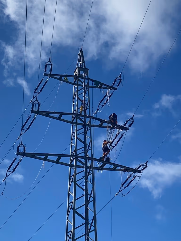 Dwóch pracowników wspina się na stalowy słup energetyczny na tle niebieskiego nieba z chmurami.