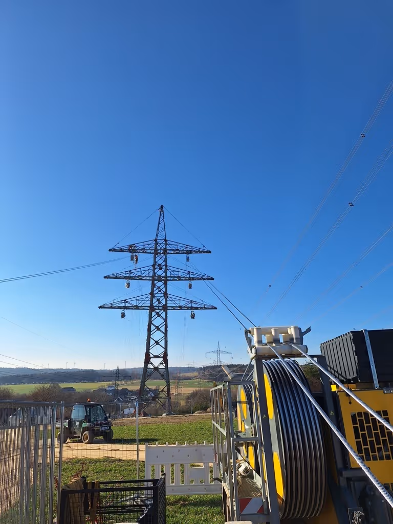 Wieża wysokiego napięcia na tle pola, z maszyną i kablami elektrycznymi na pierwszym planie w słoneczny dzień.