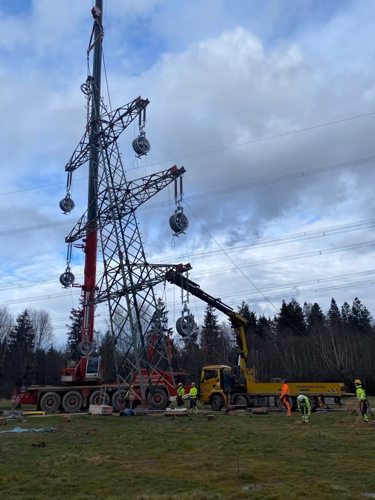 Prace konserwacyjne na linii wysokiego napięcia z żurawiami i pracownikami w odblaskowych ubraniach na zielonym polu.