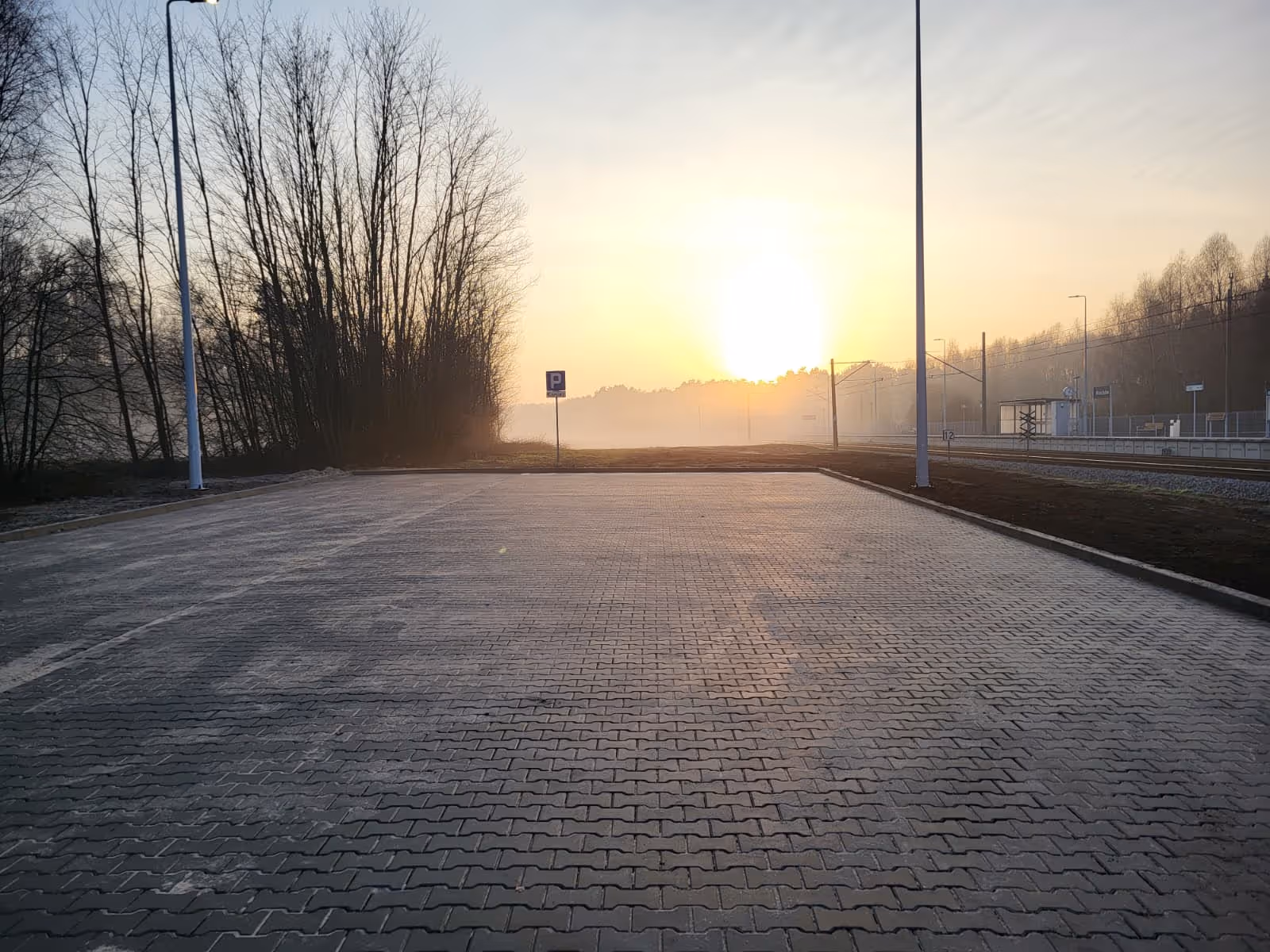 Pusty parking z kostki brukowej o wschodzie słońca z latarniami i drzewami w tle przy torach kolejowych.
