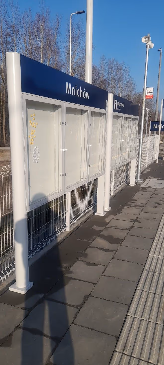 Przystanek kolejowy Mnichów z tablicą informacyjną i jasnym niebem w tle.