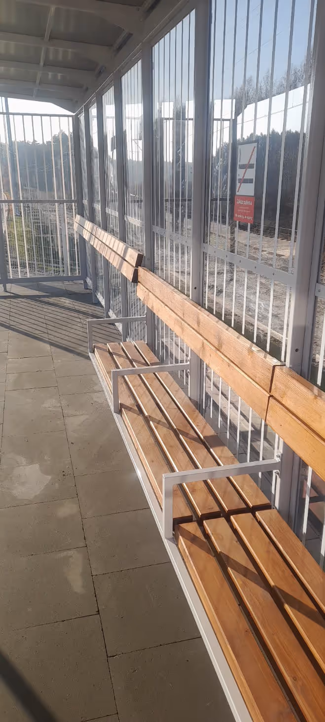 Przystanek ochroniony metalową konstrukcją i ławkami z drewnianych desek na betonowej posadzce, oświetlony promieniami słonecznymi.