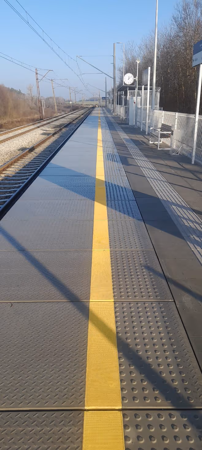 Peron kolejowy z żółtą linią ostrzegawczą i tarczą zegara na słupie oświetleniowym w słoneczny dzień.