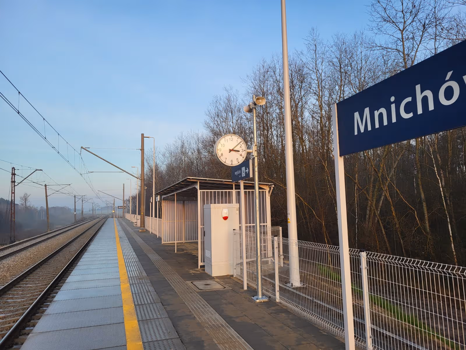 Peron stacji kolejowej Mnichów o wschodzie słońca z zegarem wskazującym godzinę 7:15.