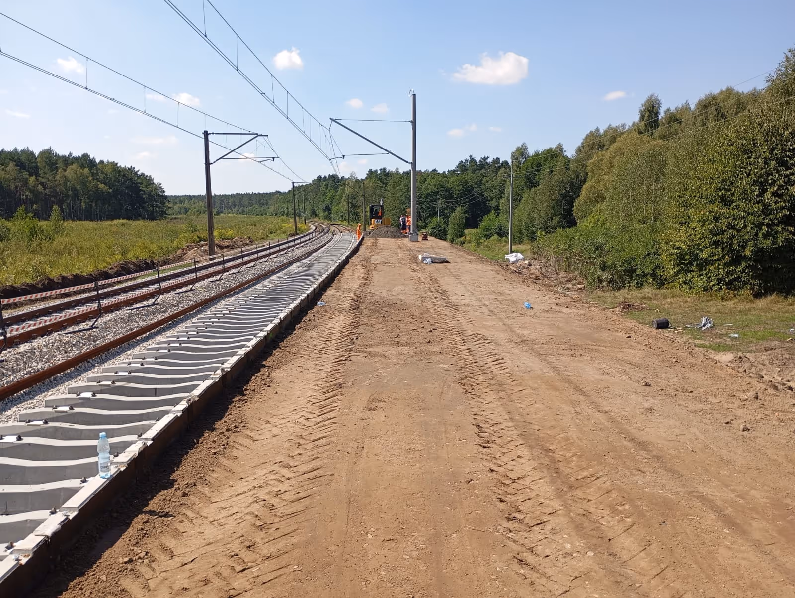 Plac budowy nowego toru kolejowego z maszyną i pracownikami na tle lasu pod pochmurnym niebem.