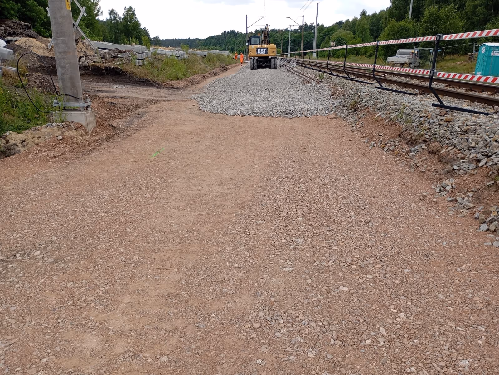 Teren budowy torów kolejowych z maszyną CAT, robotnikami i materiałami budowlanymi obok torów.