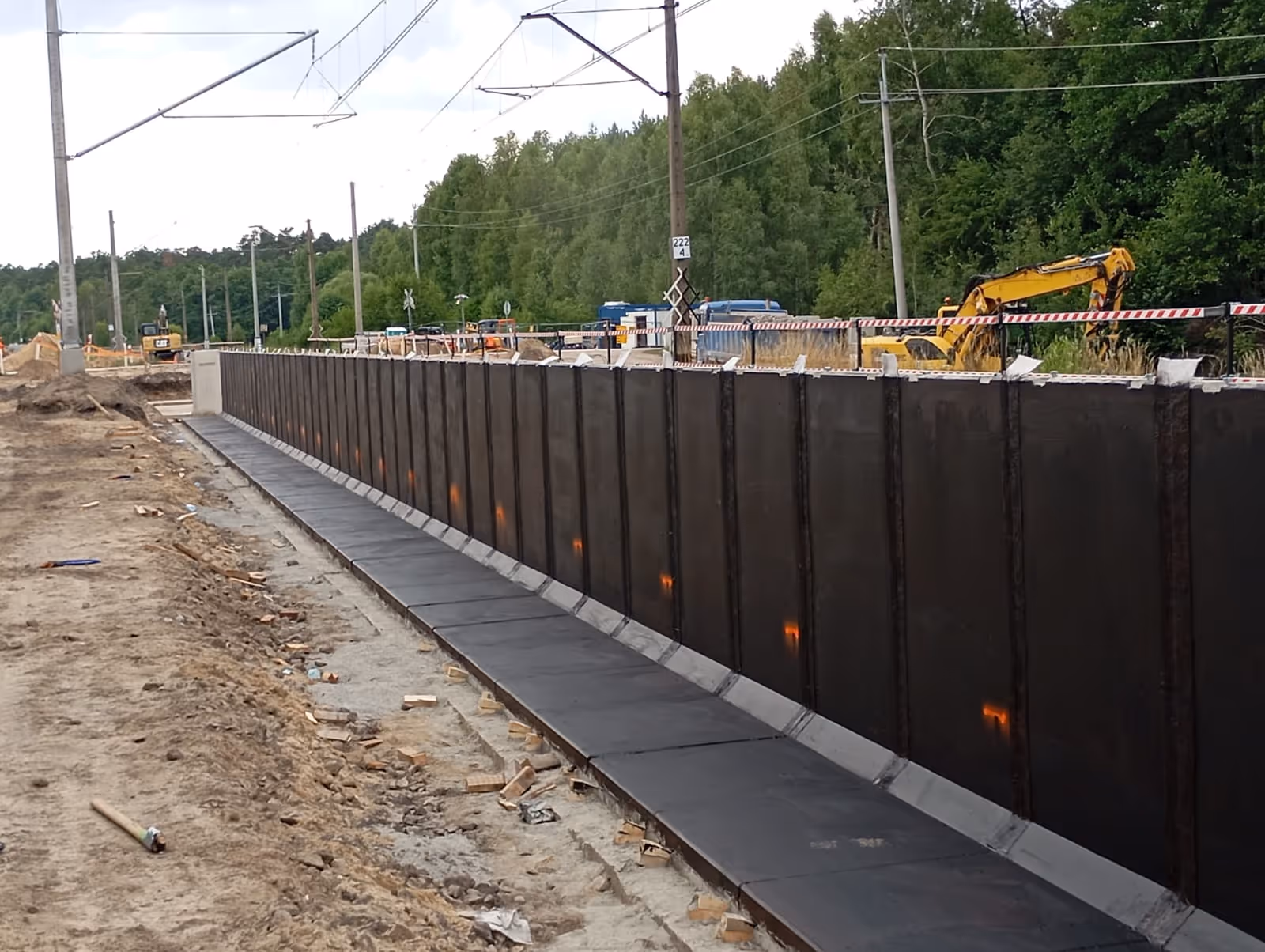 Plac budowy z wysokim metalowym murem oporowym i maszynami budowlanymi w tle przy linii kolejowej.