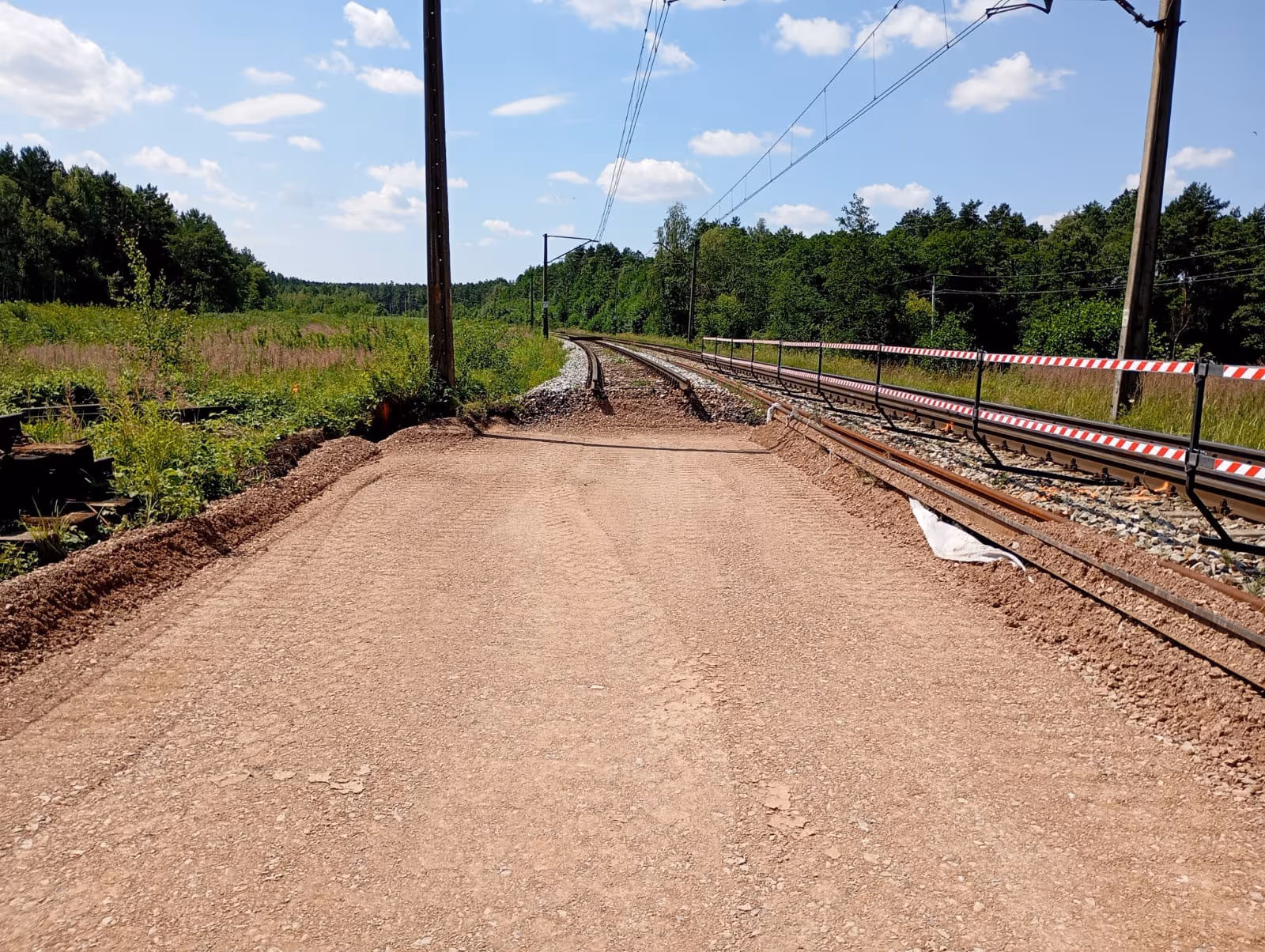 Nieułożona droga gruntowa kończąca się przed zakrętem torów kolejowych otoczona zielonymi drzewami i roślinnością.