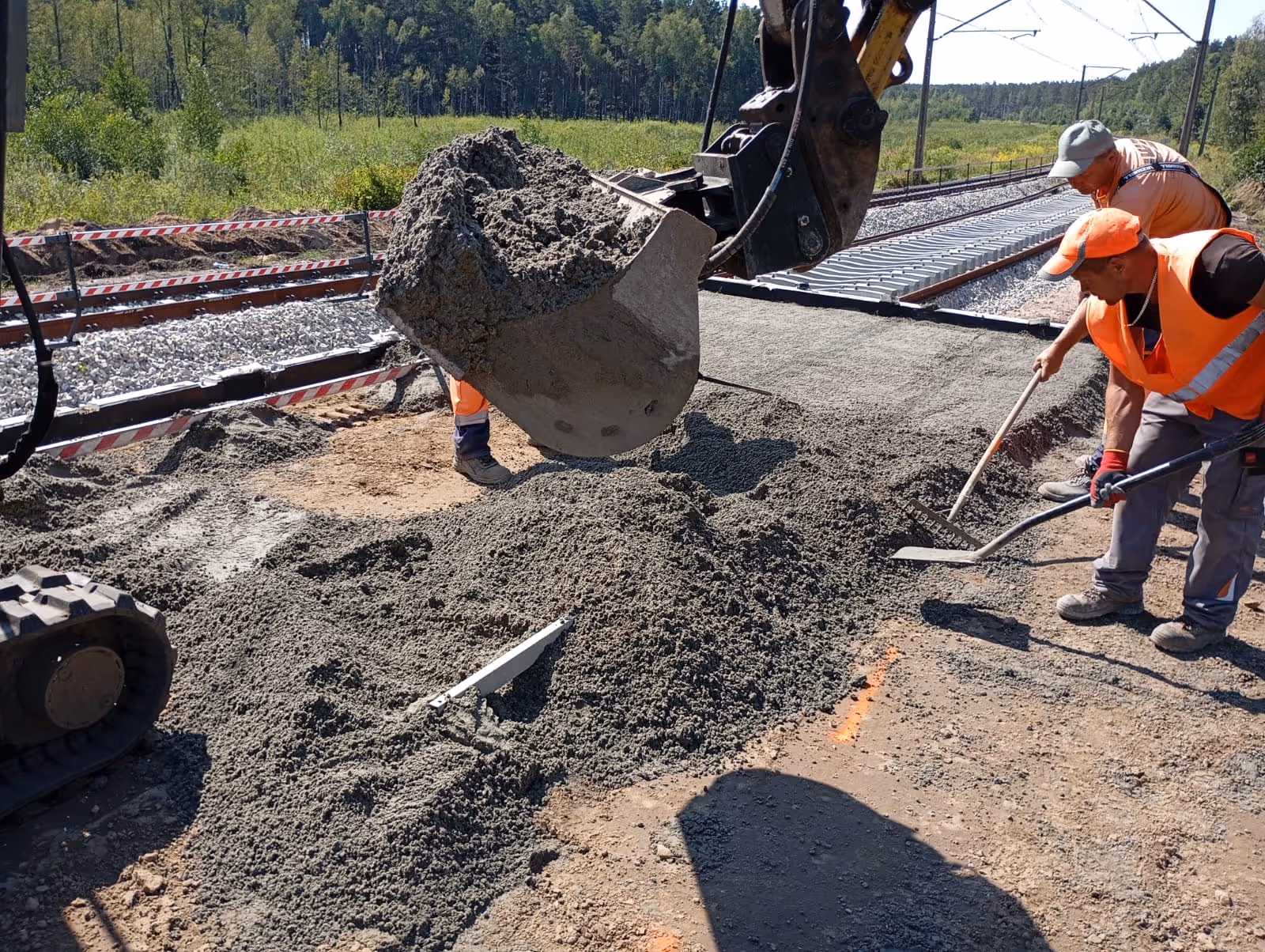 Robotnicy rozprowadzający świeży beton obok torów kolejowych w lesistym terenie.