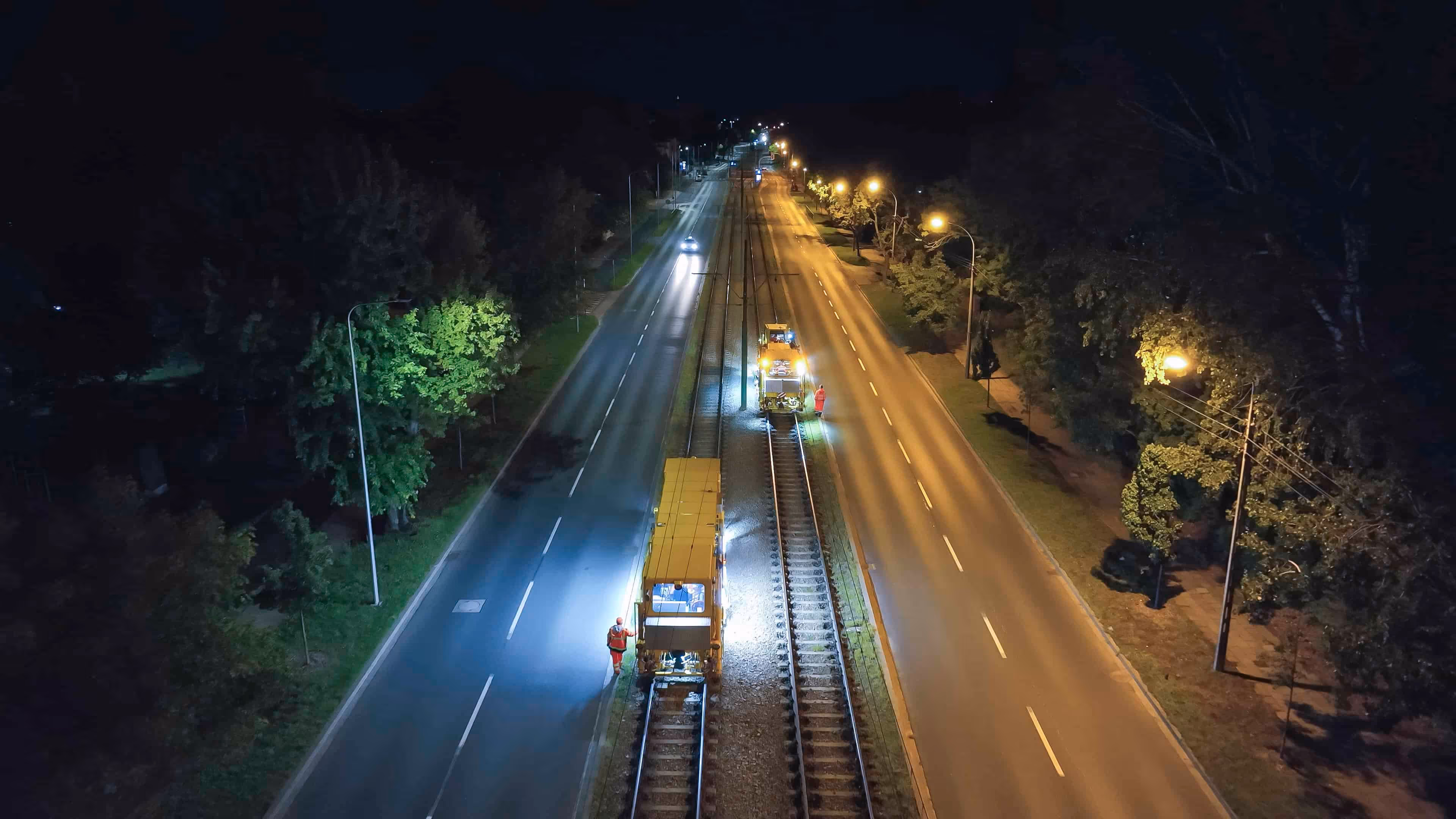 Nocna praca na torach tramwajowych między dwiema drogami oświetlonymi latarniami ulicznymi.