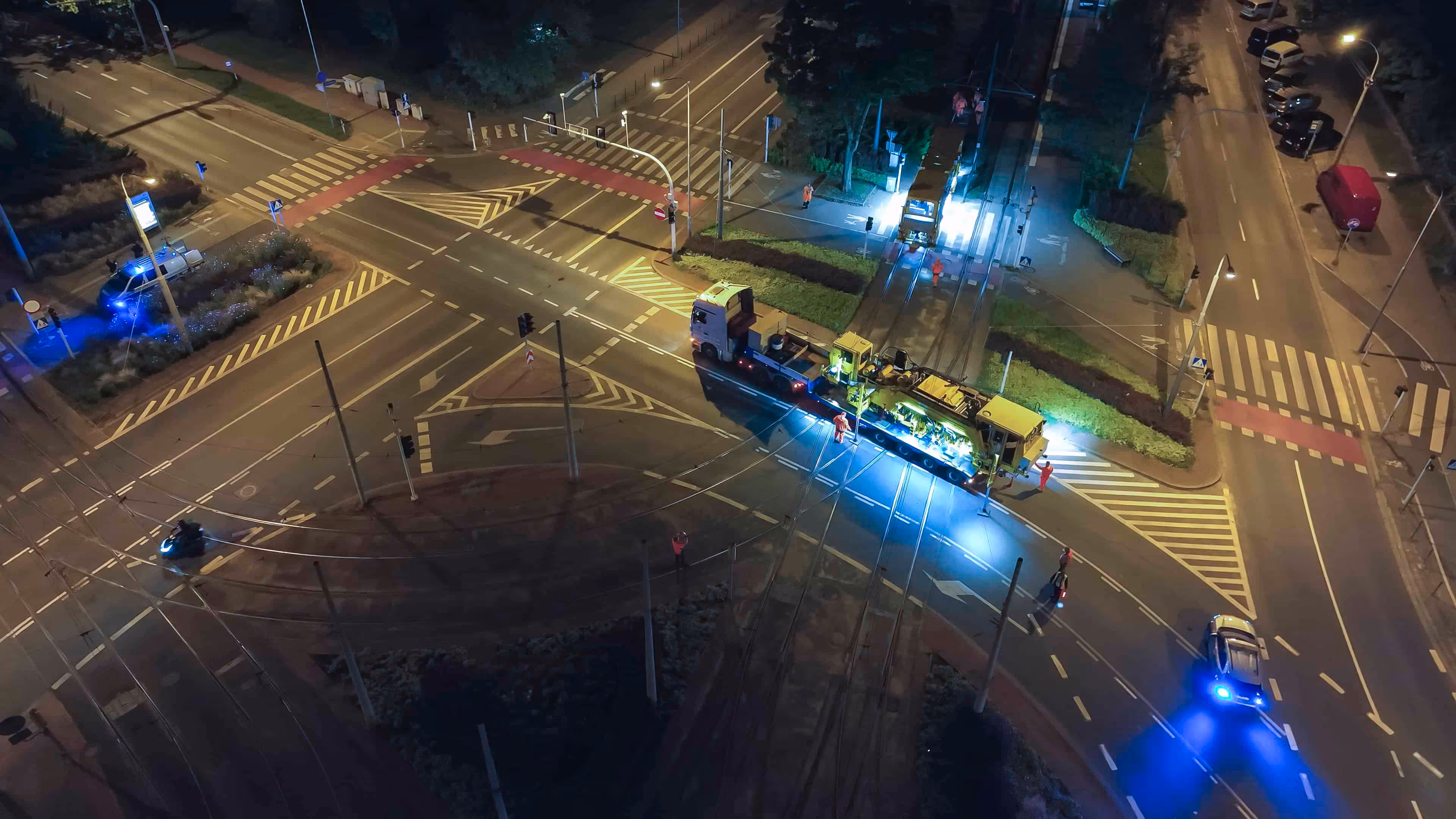 Nocna praca przy remoncie torów tramwajowych na oświetlonym skrzyżowaniu z obecnością pojazdów i pracowników.
