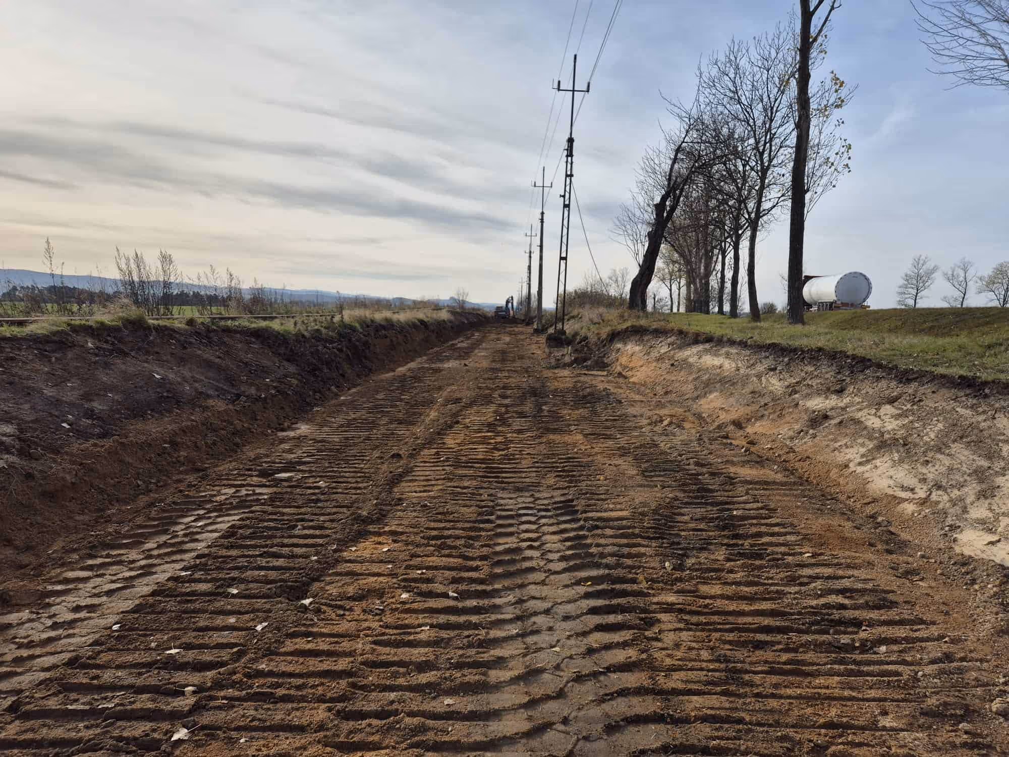 Droga gruntowa w trakcie budowy z widocznymi śladami opon maszyn budowlanych, po bokach drzewa i linia słupów energetycznych.