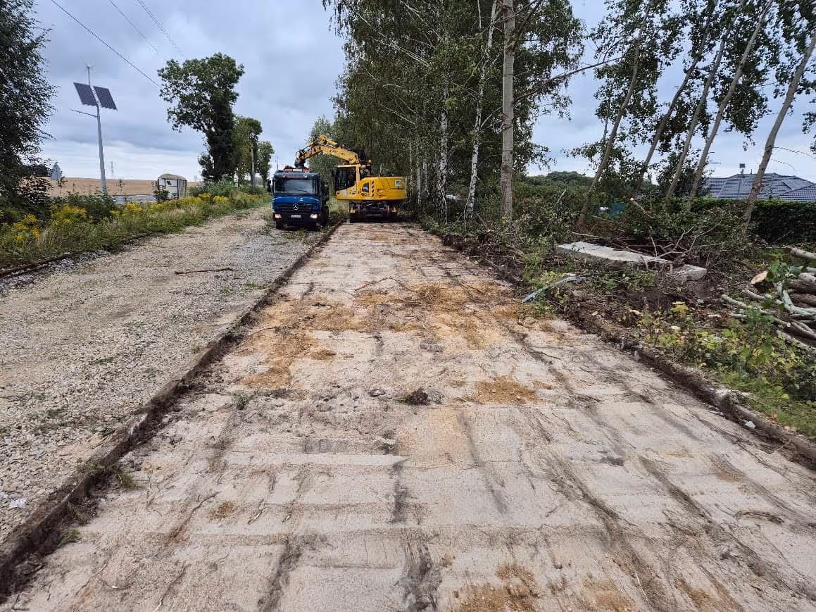 Droga w trakcie budowy między dwoma rzędami drzew, z koparką i ciężarówką stojącymi na żwirowej powierzchni.