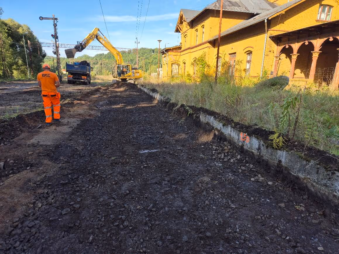 Plac budowy obok starego, żółtego budynku przy torach kolejowych z koparką i pracownikiem w pomarańczowym ubraniu.