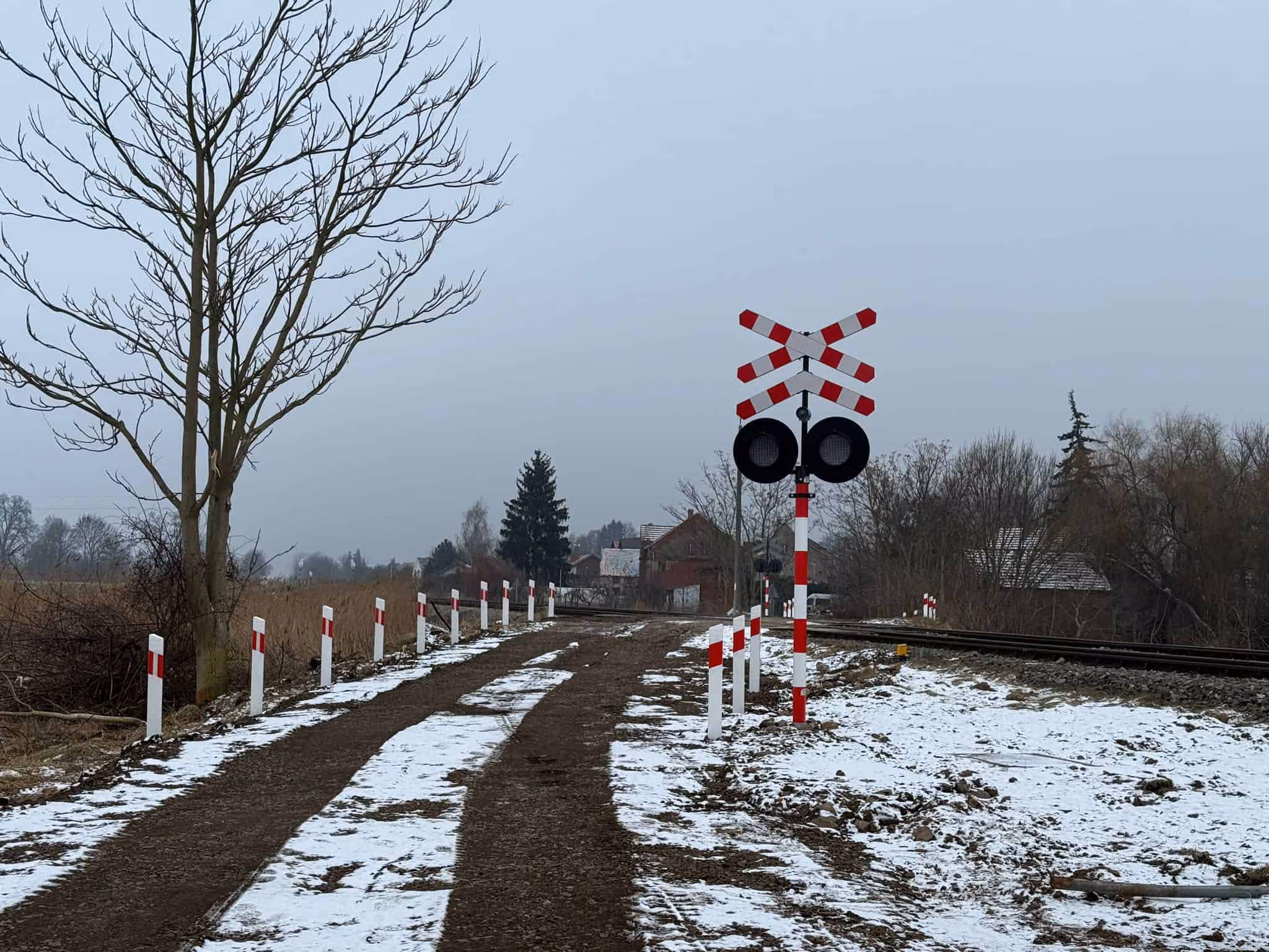Kolejowe przejście w wiejskim krajobrazie zimą z częściowo zaśnieżoną drogą i przejazdem kolejowym oznaczonym czerwono-białymi znakami.