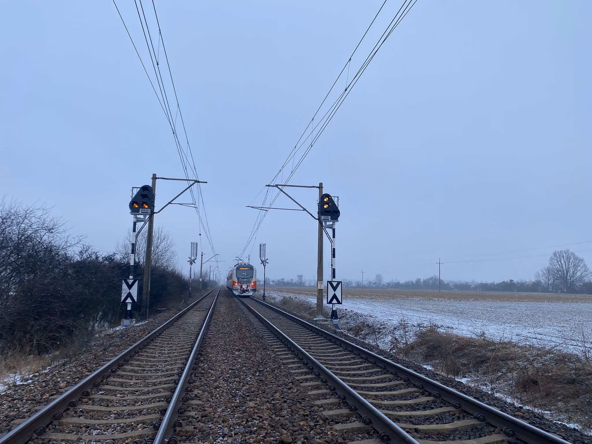 Pociąg elektryczny jedzie po torach kolejowych zimą z zapalonymi żółtymi światłami sygnalizacji.