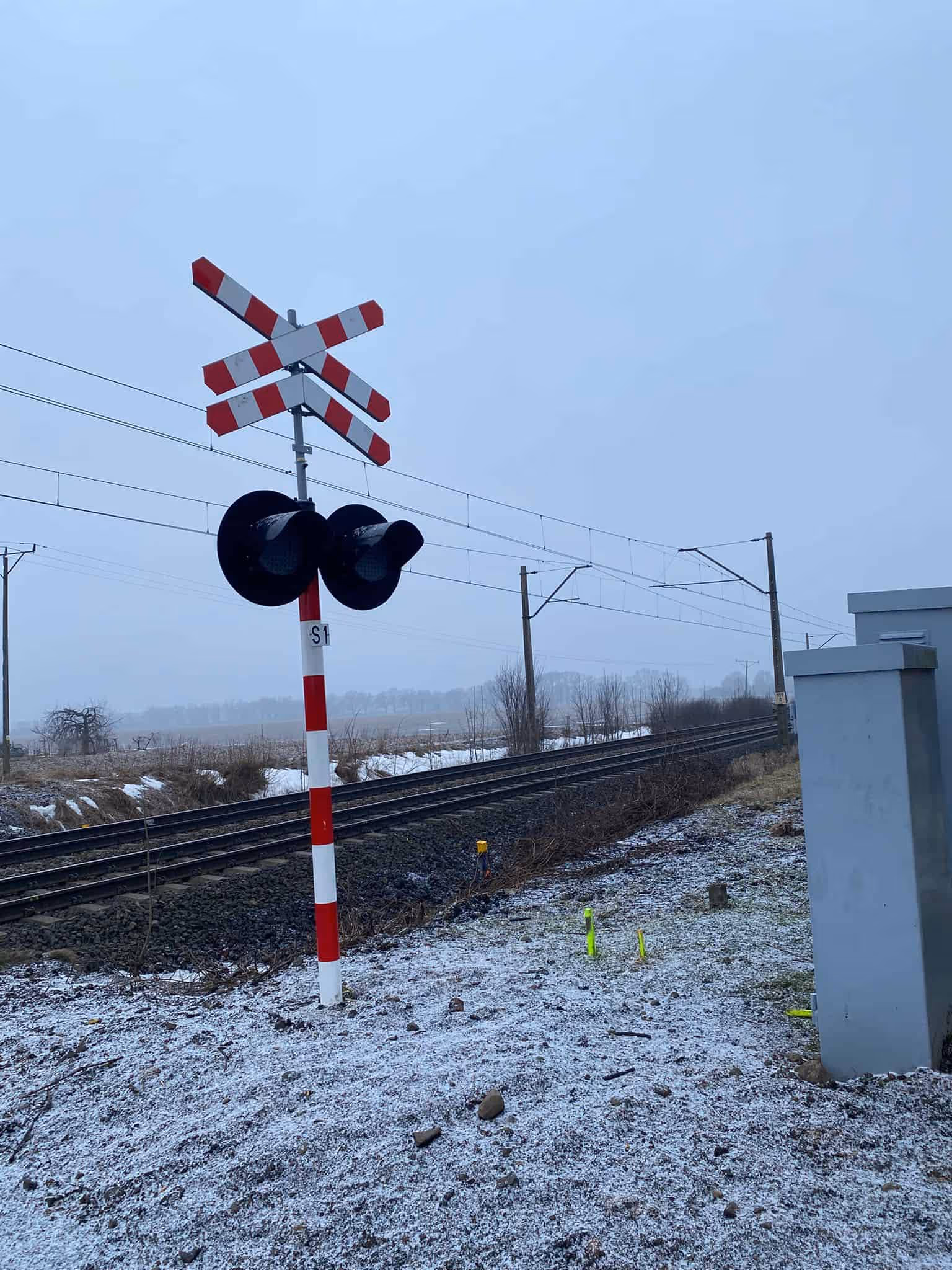 Znak ostrzegawczy przejazdu kolejowego przy torach z lekkim śniegiem i pochmurnym niebem.