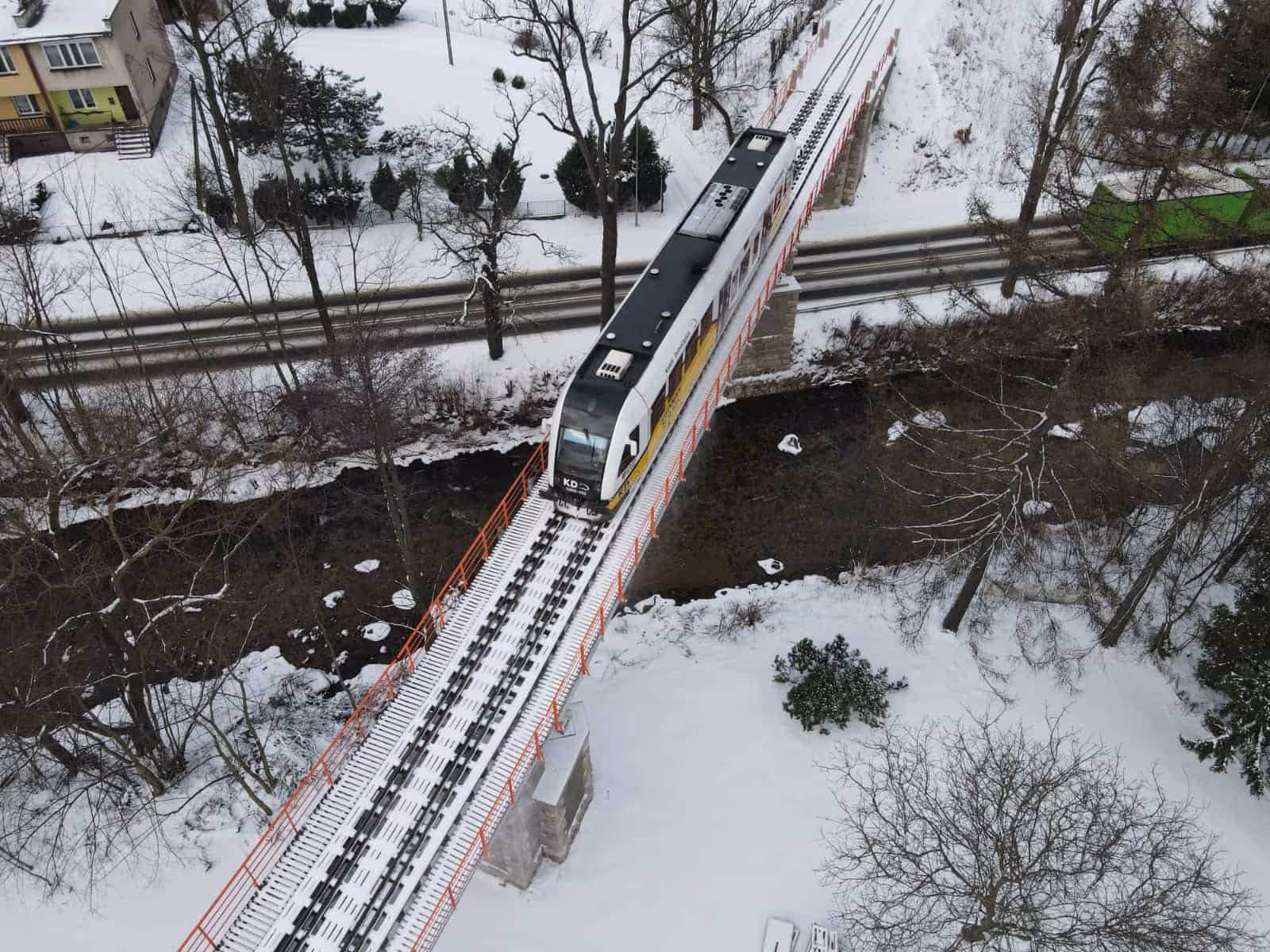 Pociąg przejeżdżający przez zaśnieżony most nad rzeką w zimowym krajobrazie.